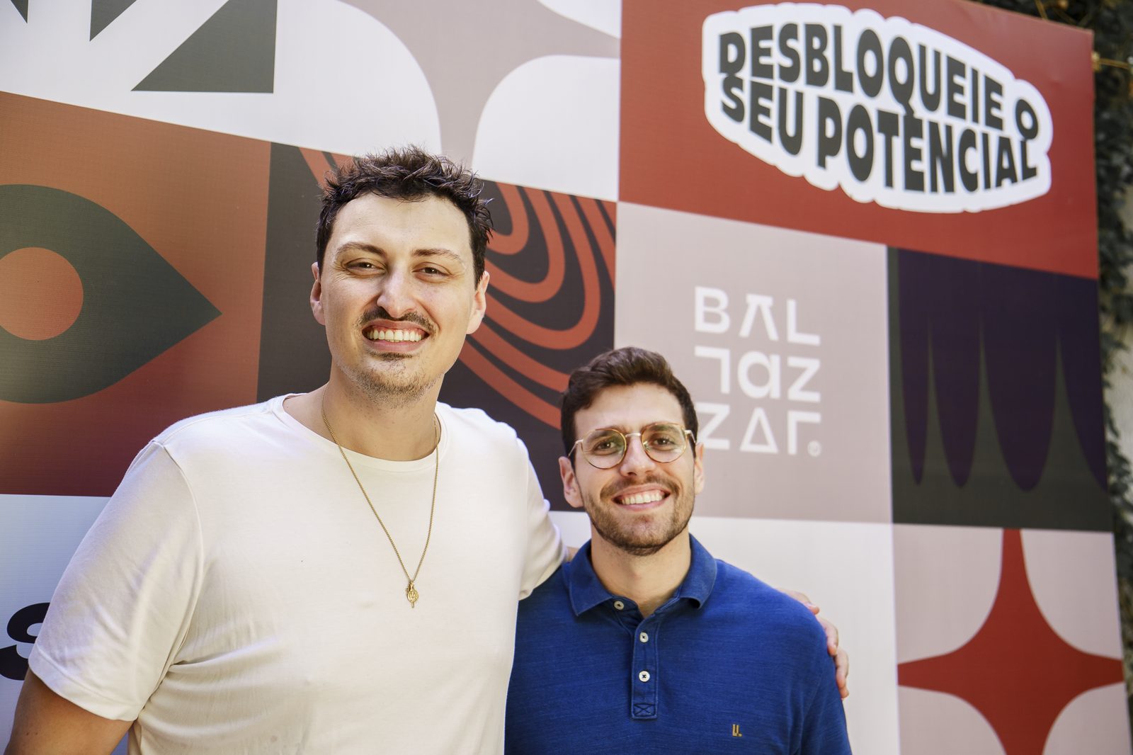 Dois convidados do Petit Comité posando juntos em frente ao backdrop da BALTAZZAR com a frase Desbloqueie o Seu Potencial